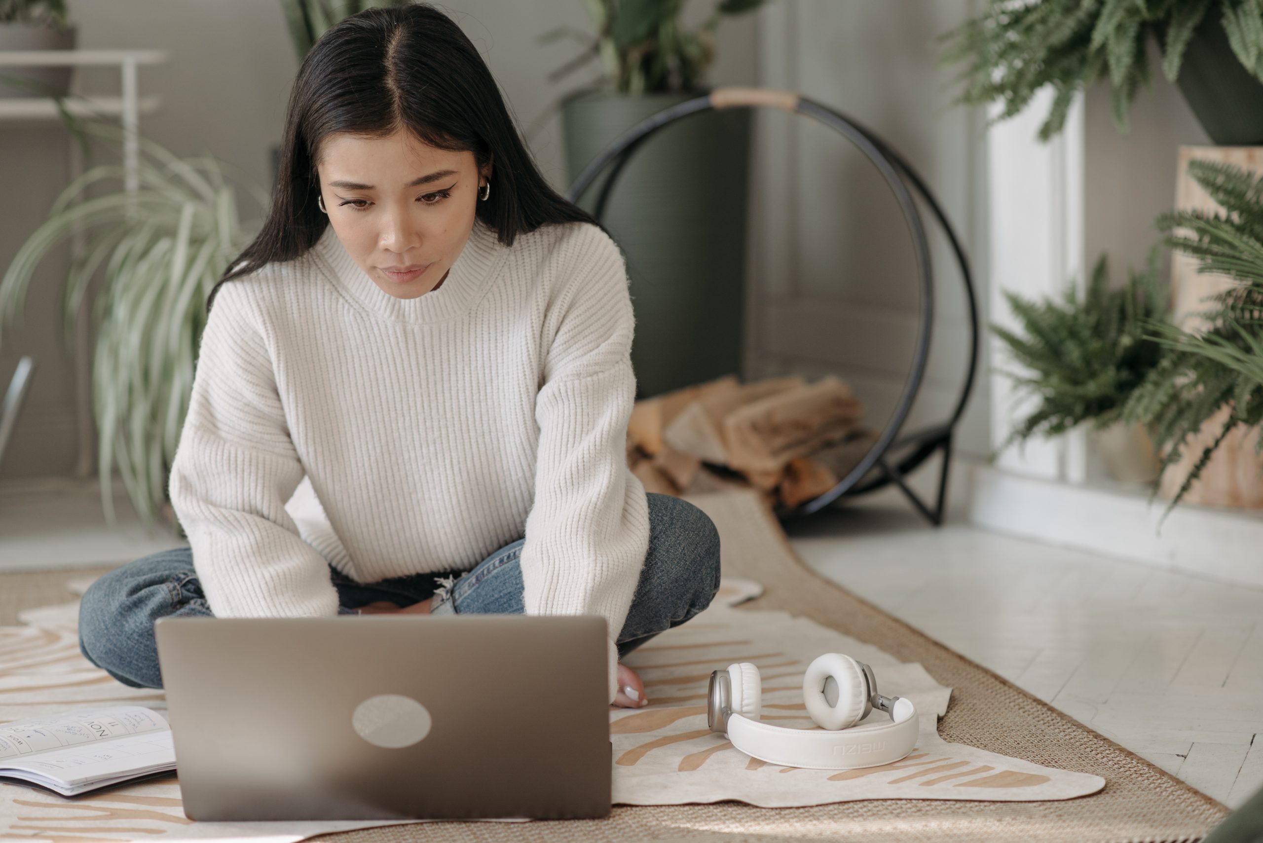 A young woman in a comfy looking jumper sits on the floor and looks at a laptop. She is concentrating but looks comfortable and like she is focusing on her task.